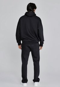 Zwart hoodie- en joggingbroekset, gemaakt van zachte stof. Hoodie heeft een grote kraag, ontspannen pasvorm en geribbelde boorden. Witte sneakers maken de look compleet.