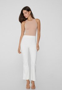 Žena s dlouhými tmavými vlasy, oblečená v tílku v béžové barvě, bílých přiléhavých kalhotách a béžových lodičkách, stojící proti běžnému světlému pozadí.