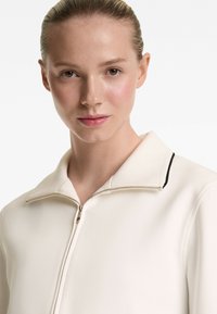 Cream zipper jacket with a collared neckline, featuring a contrasting black trim along the collar and a silver zip closure. Smooth texture.