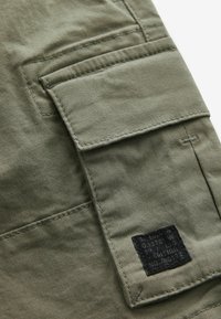 Pantaloni cargo in cotone verde oliva con una tasca applicata, cuciture a vista e un'etichetta nera con testo bianco nella parte inferiore.