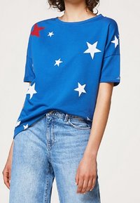 Camiseta de algodón azul con grandes patrones de estrellas en blanco y rojo. Cuello redondo y mangas cortas, combinada con jeans de un azul claro.