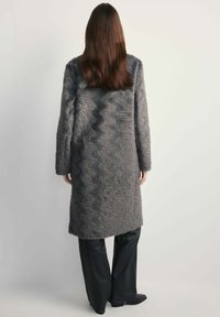 Teksturowany szary płaszcz z falistym wzorem, długimi rękawami i prostym krojem, zestawiony z czarnymi spodniami o szerokich nogawkach. Gładkie wykończenie materiału.