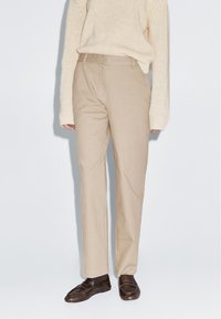 Pantalons en coton beige à coupe droite, avec une finition lisse et des passants de ceinture, associés à des mocassins en cuir noir et un pull crème.