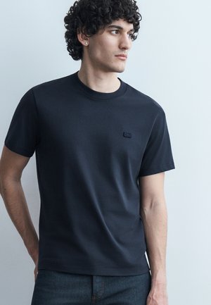 Mladý muž s kučeravými vlasmi, oblečený v jednoduchom čiernom tričku s krátkym rukávom a tmavých džínsoch, stojaci pred svetlosivým pozadím.