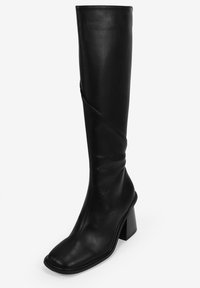 Bota de cuero negro hasta la rodilla con puntera cuadrada y tacón cuadrado. Presenta una textura suave y detalles de costura diagonal para variar el diseño.