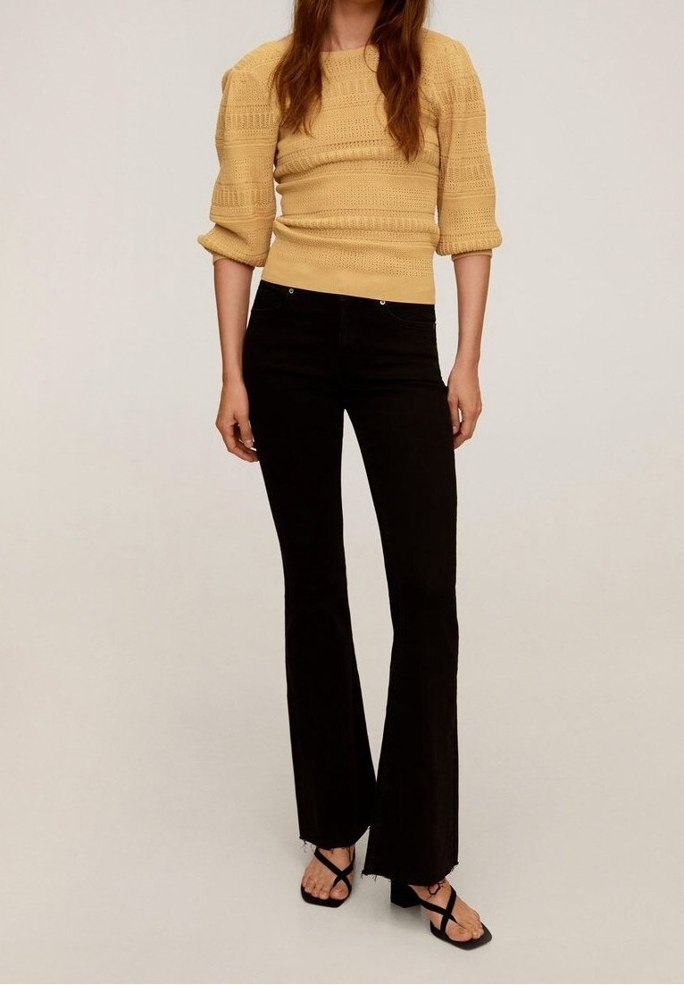 Femme portant un pull en maille jaune moutarde, un pantalon flare noir ajusté avec des ourlets effilochés, et des sandales noires à brides avec talons blocs.