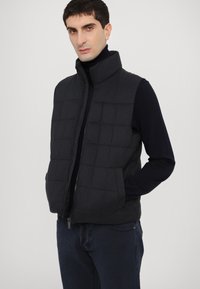 Quilted black vest with high collar and zipper front. Features side pockets and a textured fabric, worn over a dark turtleneck.