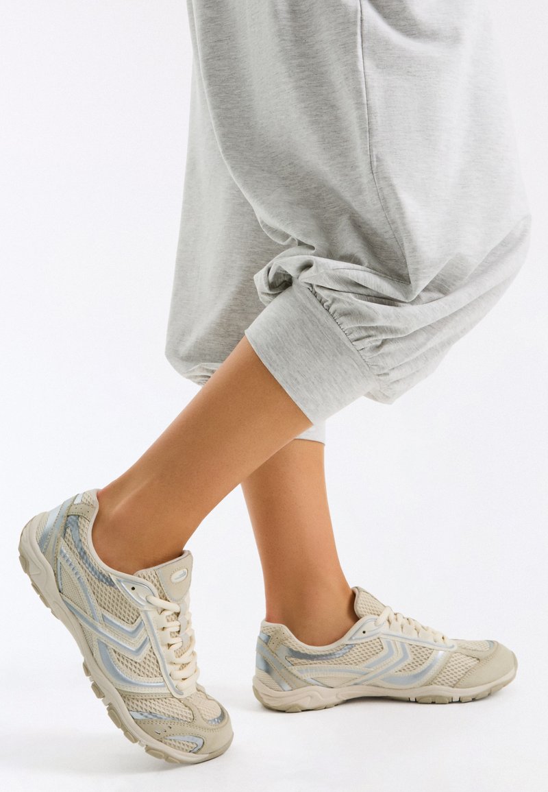 Personne portant un pantalon de survêtement gris clair roulé au-dessus des chevilles et des baskets en mesh beige avec des détails blancs et bleus sur fond blanc.