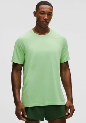 Ung mand iført en lys grøn kortærmet skjorte og mørkegrønne shorts, der står foran en ensfarvet grå baggrund.