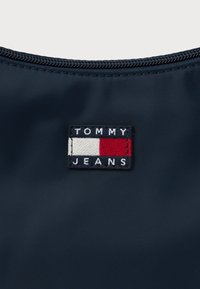 Prim-plan al unui material albastru marin cu un fermoar deasupra și o etichetă cu logo-ul Tommy Jeans, care include culorile alb, roșu și albastru marin, centrată sub fermoar.
