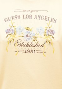 Κίτρινο ύφασμα με το κείμενο «GUESS LOS ANGELES Established 1981» και σχέδιο με λουλούδια που περιλαμβάνει ανοιχτό μπλε και μωβ λουλούδια με κίτρινα φύλλα.