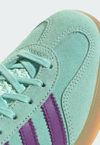 Mintgrön mockaskor med en texturerad yta, lila randiga accentdetaljer och en ljusbrun gummisula med vita sömmar.