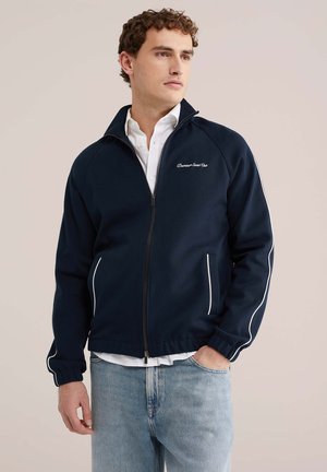 Mees kannab tumesinist lukuga jopet valge toruga ja "Downtown Social Club" logoga, valget särki ning helerohelisi teksaseid beežitaustal.