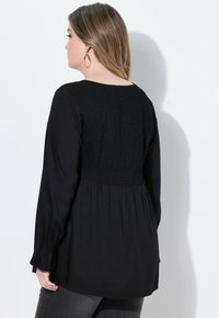 Schwarze Bluse mit langen Ärmeln, die eine Raffung am Rücken, einen runden Halsausschnitt und eine geraffte Textur aufweist. Weicher Stoff mit lässiger Passform.