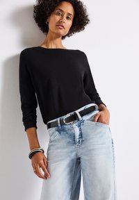 Svart långärmad topp i kombination med ljusblå vida denimjeans, med ett svart bälte och lager av cirkulära armband. Slät textur.