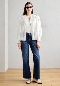 White pleated oversized shirt, fitted white top, dark blue flared jeans, cream shoes with studs, and black sunglasses. Wood floor background.
