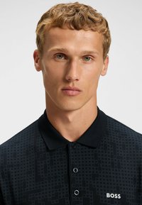 Svart polo skjorta med en texturerad design och geometriska mönster, en klassisk krage och tre knappar. "BOSS"-logotypen visas på bröstet.