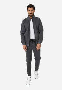 Giacca sportiva grigia con colletto alto e zip, abbinata a pantaloni jogger coordinati e sneakers bianche. Il tessuto ha una consistenza morbida e una vestibilità rilassata.