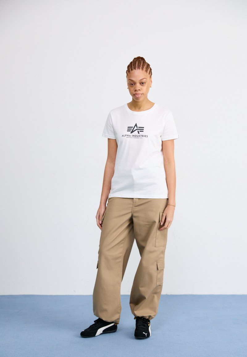 T-shirt blanche avec le logo « Alpha Industries » en noir, associée à un pantalon cargo beige ample et des baskets noires, sur un fond simple.