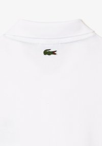 Camiseta polo blanca con un cuello clásico y un logo de cocodrilo bordado en verde en la parte posterior, cerca del cuello.
