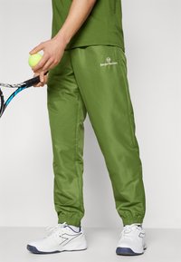 Zelene, lagane atletske hlače s elastičnim manžetama i logotipom sa strane, uparene s bijelim sportskim cipelama i lopticom za tenis.