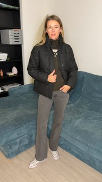 Black puffer jacket with a zip, worn over a dark green sweater. Gray flared jeans and white sneakers. Background features a blue corduroy couch.