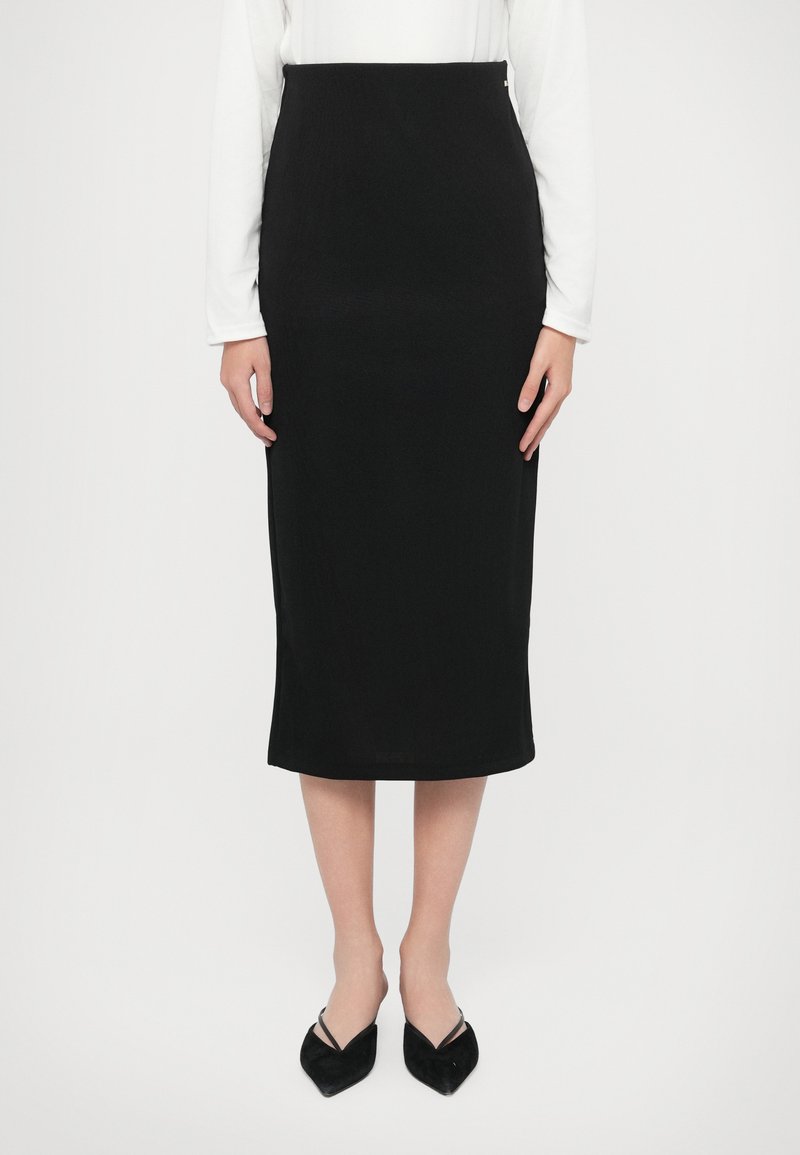 Μαύρη πλεκτή μολυβοθήκη με ψηλή μέση και μήκος μέχρι το γόνατο, κατασκευασμένη από υφασμάτινο υλικό με υφή, σε συνδυασμό με μυτερές μαύρες παπούτσια.