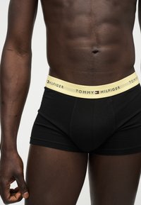 Αρσενικός κορμός και άνω πόδια που φορούν μαύρα μποξεράκια με ανοιχτό κίτρινη μέση με λογότυπο Tommy Hilfiger, στέκονται σε ανοιχτό φόντο.