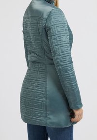 Chaqueta acolchada en color azul verdoso con un alto cuello, que presenta un patrón geométrico en la parte posterior y las mangas. Fabricada con un material suave y brillante.