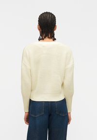 Personne aux cheveux tressés portant un pull en maille crème et un jean bleu foncé, debout de dos sur un fond blanc uni.