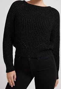 Pull noir côtelé avec un col bateau, manches longues et des détails découpés à la taille, fabriqué dans un matériau texturé et scintillant.