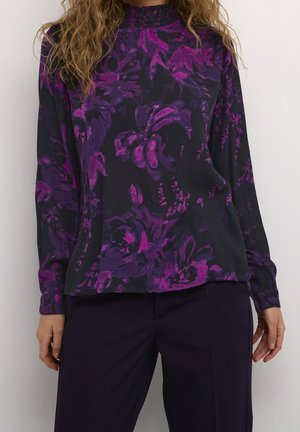 Vrouw draagt een zwart shirt met lange mouwen en paarse bloemenpatronen, gecombineerd met een donkere broek, tegen een effen witte achtergrond.