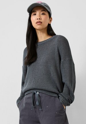 Junge Frau trägt einen grauen Strickpullover, dunkle Hosen mit Kordelzug und eine graue Baseballkappe mit einem pinken Aufnäher, steht mit den Händen in den Taschen.