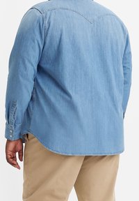 Mężczyzna w jasnoniebieskiej koszuli denimowej i beżowych spodniach, widoczny z tyłu, z lewą ręką swobodnie opartą na boku.