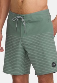 Bañador verde hecho de tela suave con rayas horizontales grises, cinturilla elástica y cordón, con un logo redondo de RVCA en el dobladillo.