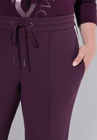 Paars broek met trekkoord in de taille, voorzien van zijzakken en witte zijstrepen, gedragen met een bijpassend paars topje, met een hand in de zak.