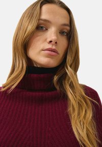 Burgundy stickad polokrage med ribbat mönster. Kragen är hög och åtsittande, i kontrast med ett svart innerlager.