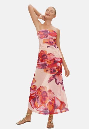 Kobieta w różowej, bezramiennej sukni maxi z dużym pomarańczowo-fioletowym kwiatowym wzorem, stojąca z jedną uniesioną ręką, nosząca sandały z kwiatowymi detalami.