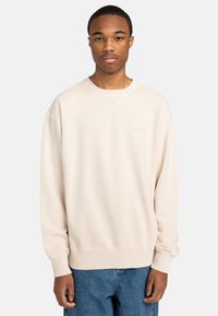 Sudadera color crema con puños y dobladillo acanalados. Presenta un escote redondo y un logo sutil en el pecho. Llevada con jeans de denim azul.