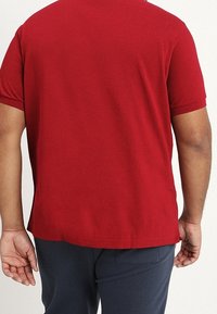 T-shirt rouge à manches courtes en texture lisse et tissée, de coupe droite avec un ourlet légèrement arrondi. Porté avec un pantalon bleu marine.