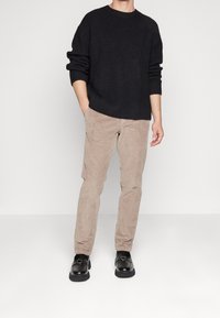 Svart stickad tröja, beige kordflöjlsbyxor och svarta skor. Tröjan har en avslappnad passform och ribbat mönster; byxorna har ett mjukt, lätt upphöjt mönster.