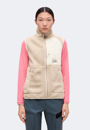 Persoon draagt een beige fleecevest met crème borstzak over een roze shirt met lange mouwen en een donkerblauwe broek, staand tegen een lichte achtergrond.