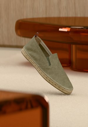 Olívazöld vászon espadrille fonott juta talppal és trikolor húzófüllel, bézs szőnyeg és áttetsző borostyán színű bútorok felett lebegve.