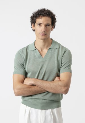 Hombre con pelo rizado y oscuro que lleva una camisa polo de punto de manga corta color salvia y pantalones blancos, de pie con los brazos cruzados sobre un fondo blanco.