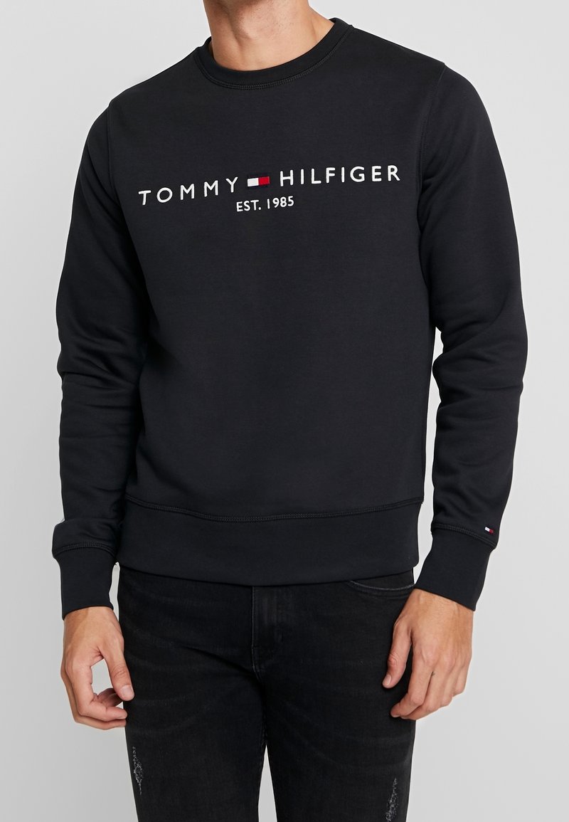 Homme portant un sweat-shirt noir Tommy Hilfiger avec logo et texte "EST. 1985", associé à un jean noir sur fond gris.