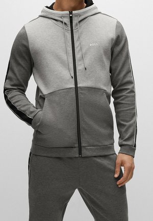 Homme portant un sweat à capuche zippé bicolore gris avec logo "BOSS" et pantalon de survêtement gris assorti, mains dans les poches, posture détendue.