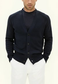 Mörkblå kardigan med V-ringning, knappstängning fram, ribbade muddar och nederkant, tillverkad av ett slätt stickat material, bärs över en mörk skjorta.