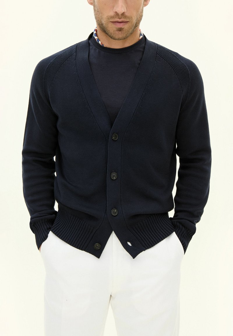Mörkblå kardigan med V-ringning, knappstängning fram, ribbade muddar och nederkant, tillverkad av ett slätt stickat material, bärs över en mörk skjorta.