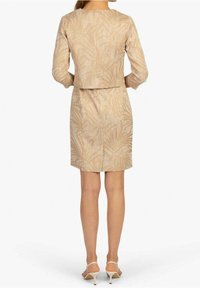 Beige kort klänning med en figurnära liv och utsvängd fåll, prydd med ett subtilt palmbladsmönster och trekvartsärmar. Skorna är ljusa klackar.