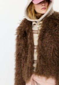 Kvinde iført en blød brun jakke over en stribet hættetrøje, pink bukser og en pink kasket, der kigger til siden mod en ensfarvet baggrund.